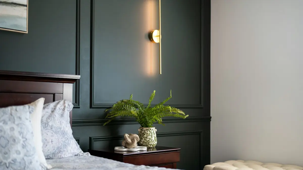 A cozy bedroom corner featuring a bedside table, a plant, and modern wall lighting.