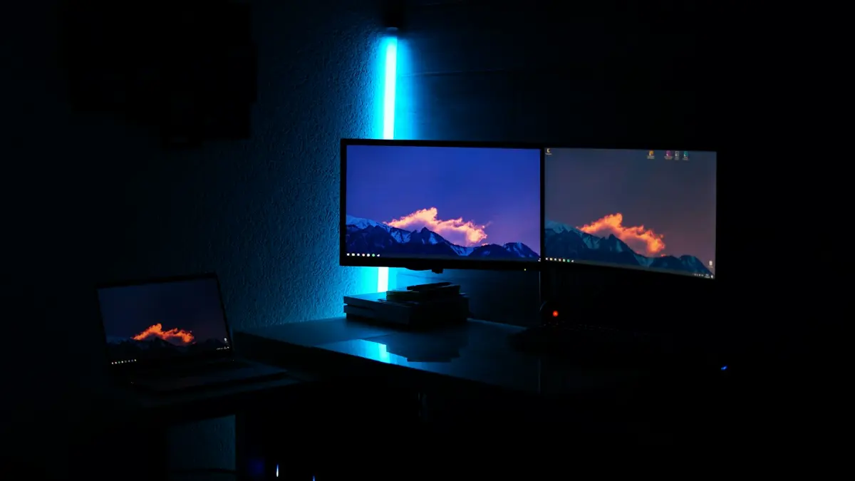 A dimly lit room with two computer monitors and a laptop, illuminated by a blue light strip.