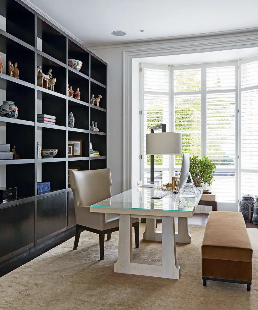 A bright home office featuring a glass-top desk, comfortable chair, and bookshelf with decorative items.